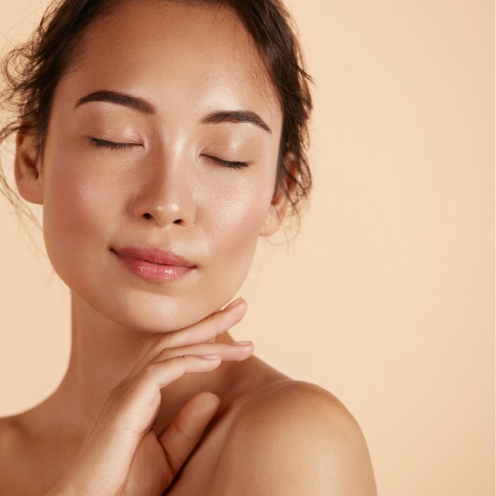 Beautiful asian girl model touching fresh glowing hydrated facial skin on beige background closeup. Skin care concept