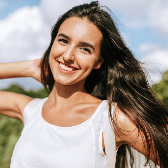 Happy young brunette woman smiling broadly with a windy blowing long hair in the park, posing on nature background.