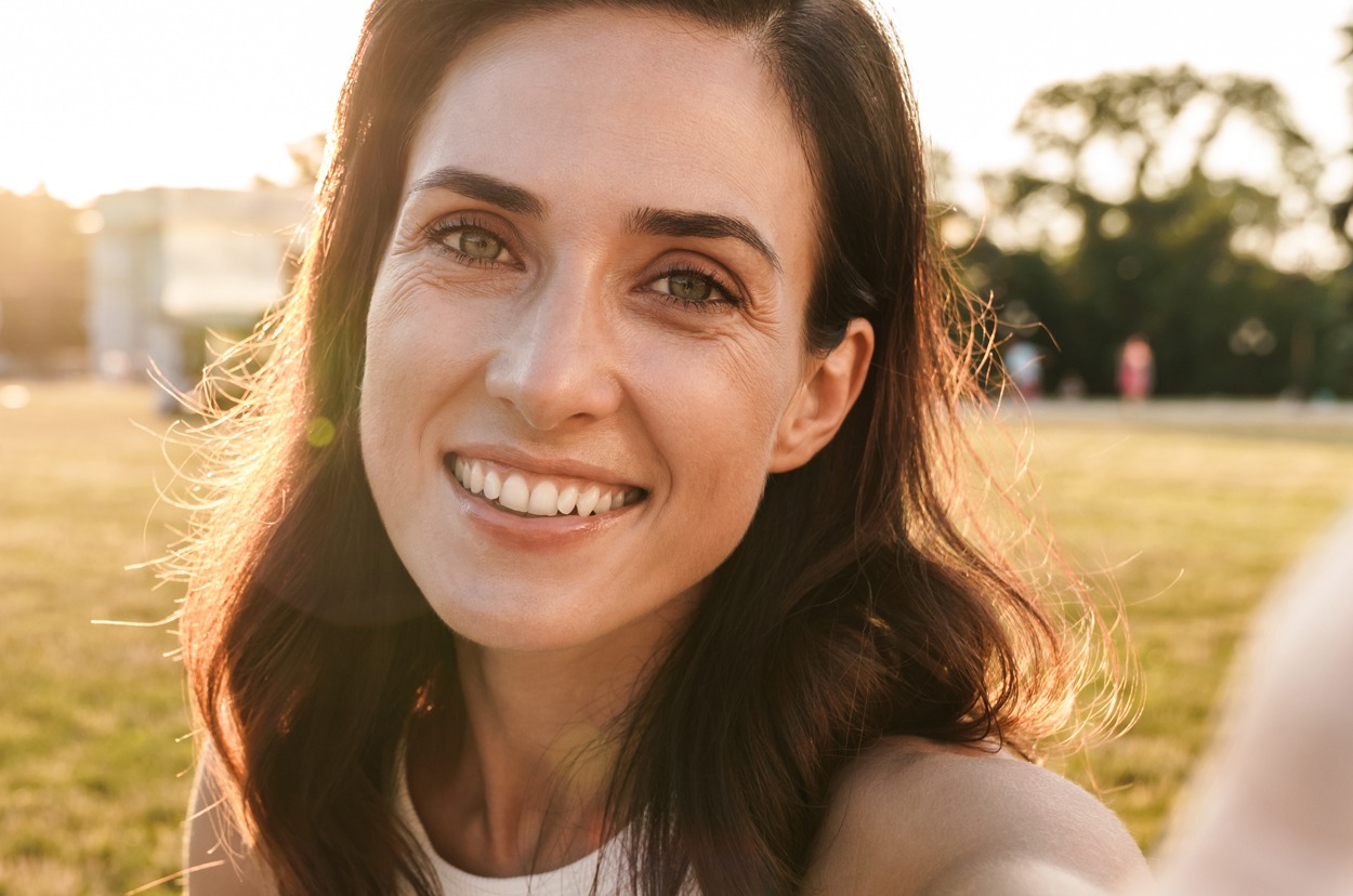 Image of smiling middle-aged woman taking selfie photo while sitting on grass in summer park