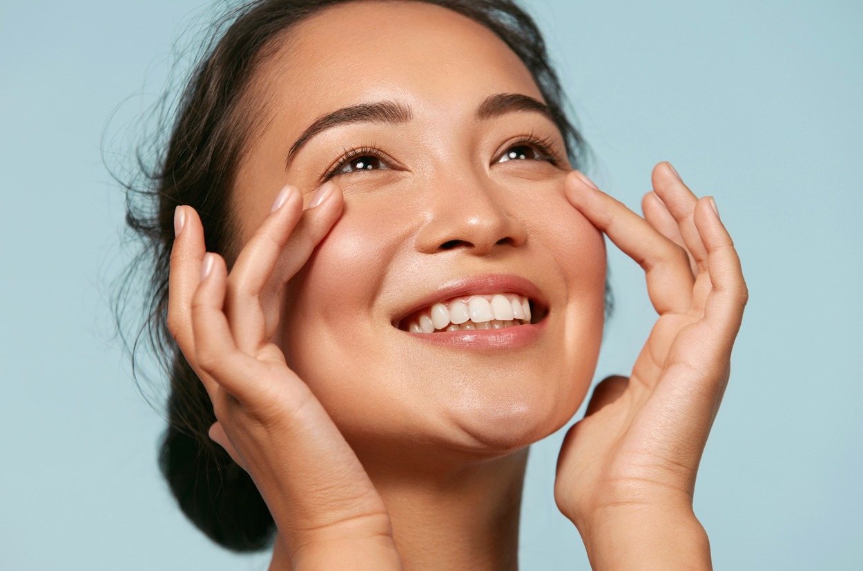 Skin care. Woman with beauty face touching healthy facial skin portrait. Beautiful smiling asian girl model with natural makeup touching glowing hydrated skin on blue background closeup