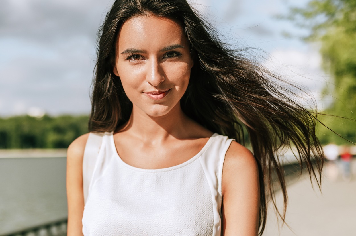 Beautiful young brunette woman smiling broadly with a windy blowing long hair in the park on a background with a lake.