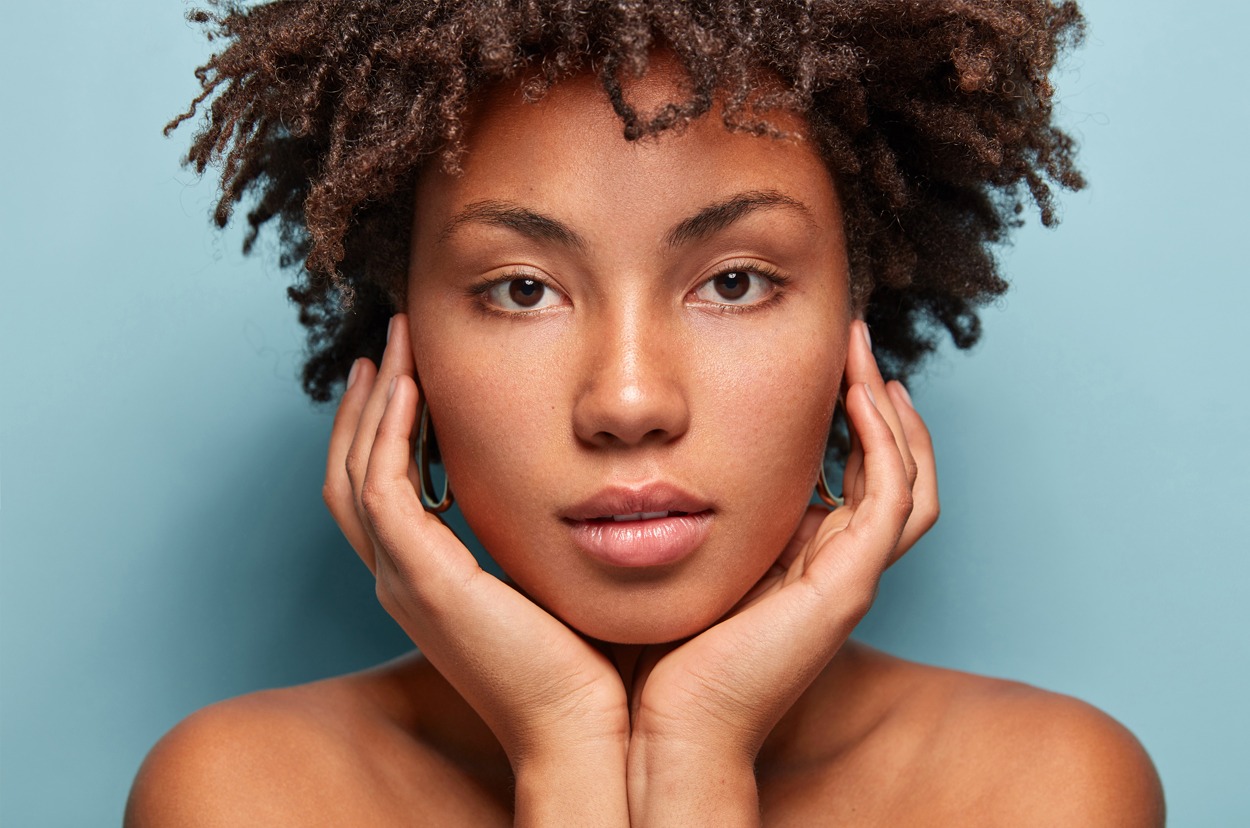 Close up shot of black young woman with Afro hairstyle, touches cheeks with both hands, has bare shoulders, tender look isolated over blue background, looks directly at camera.