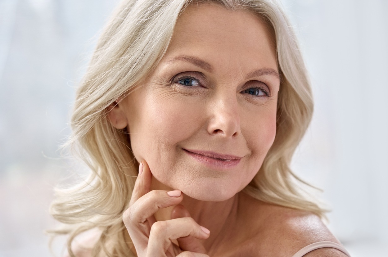 Smiling happy attractive 50s middle aged mature blond woman, old lady looking at camera advertising anti age face skin and body care treatment cosmetics posing in bathroom.