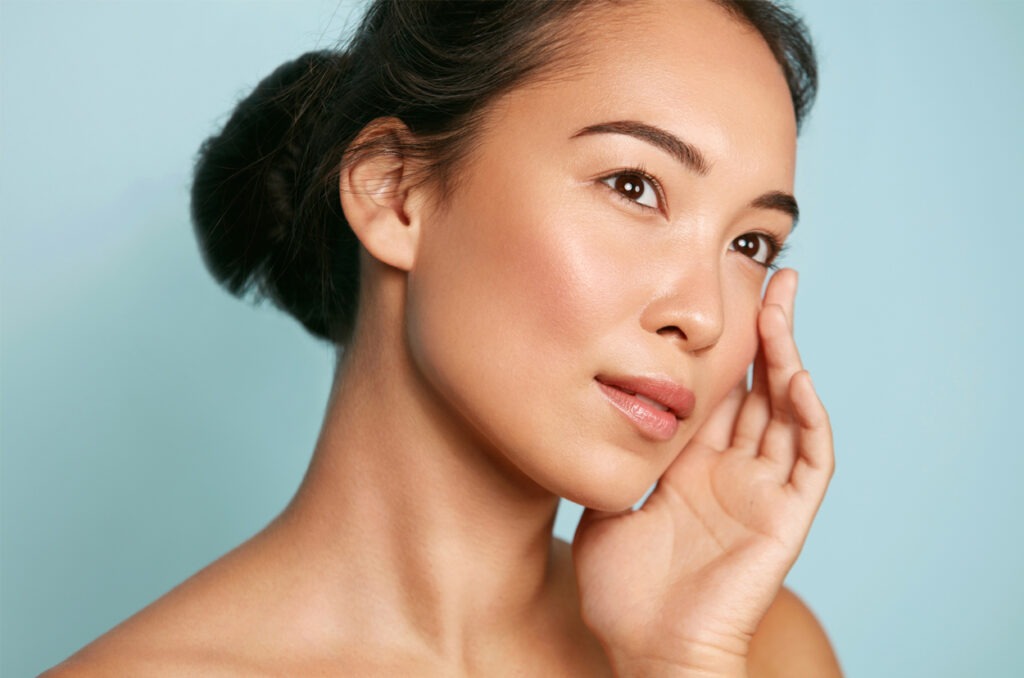 Skin care. Woman with beauty face and healthy facial skin portrait. Beautiful asian girl model with natural makeup touching glowing hydrated skin on blue background closeup.